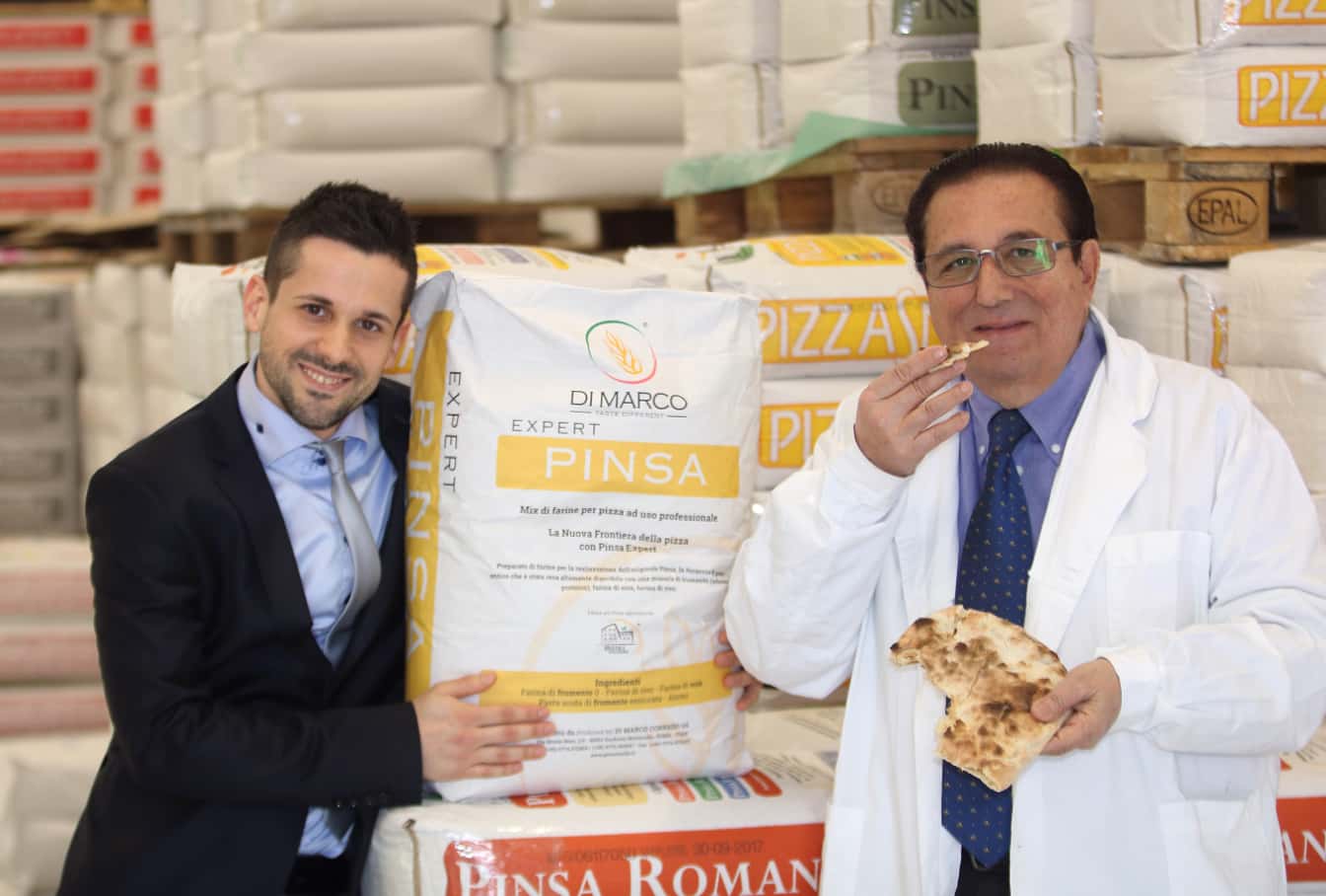 Foto di Corrado di Marco, con un membro dello staff, mentre assaggia un pezzo di Pinsa con dei sacchi di farina alle spalle.
