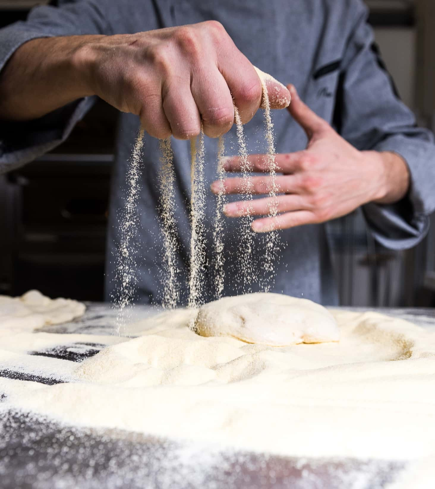 Dettaglio della mano di un cuoco che sparge la farina sul piano di lavoro durante l’impasto di una Pinsa.