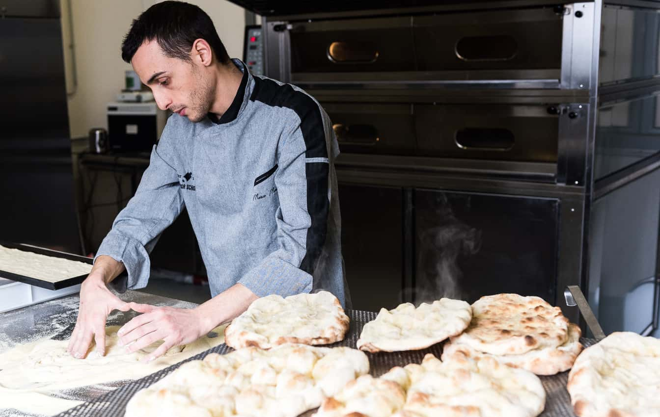 Foto di un cuoco mentre impasta delle pinse in una cucina professionale e con numerosi impasti pronti sul tavolo.