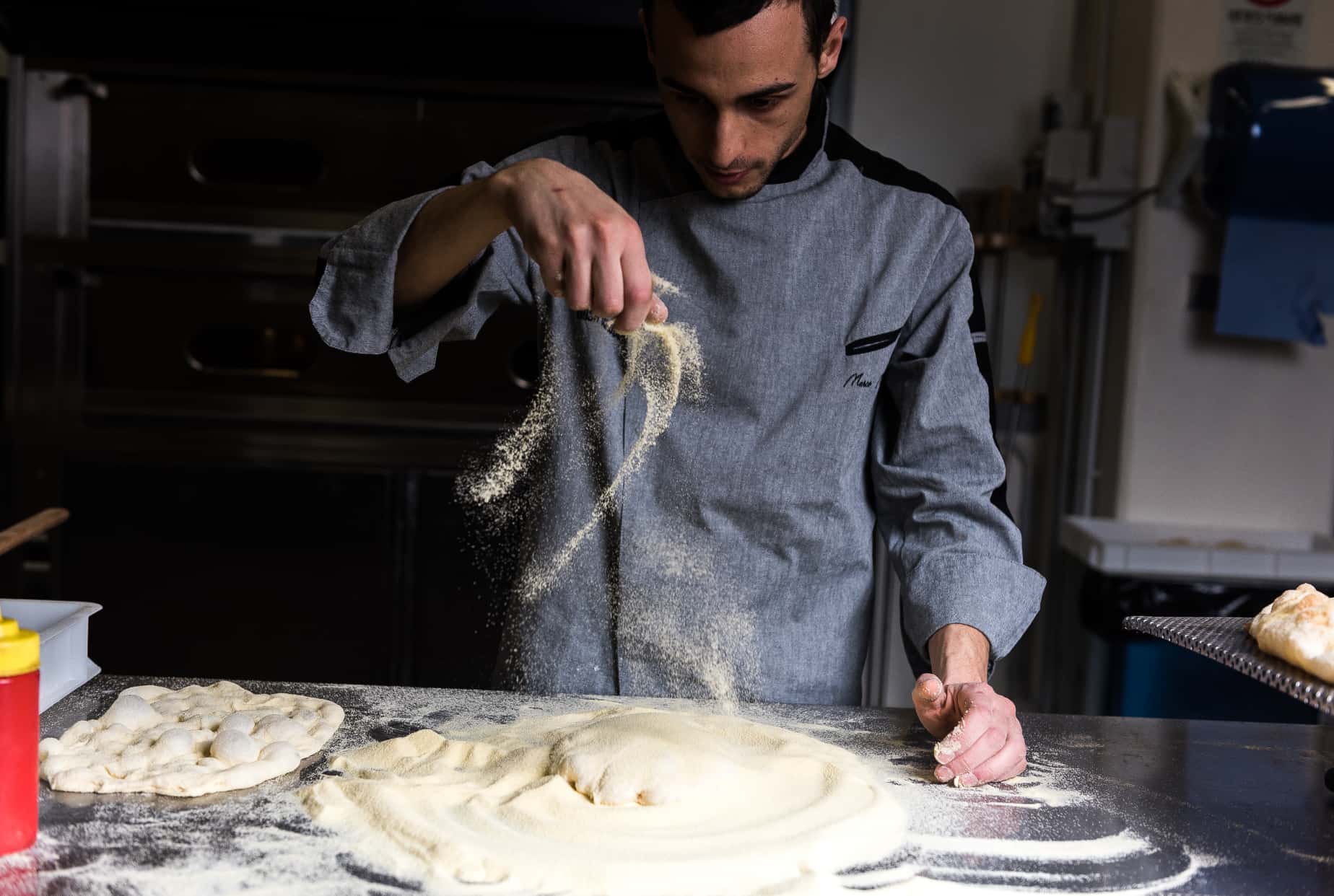 Foto di un cuoco in divisa che sparge della farina su un impasto di Pinsa Romana.
