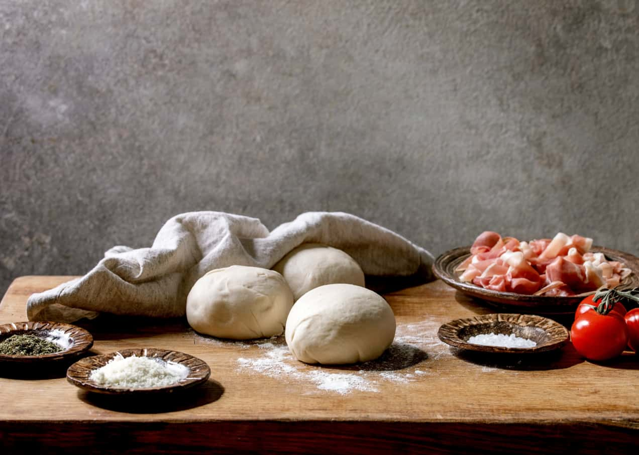 Photo of dough balls on a wooden table and with plates with salt, ham, cherry tomatoes next to them