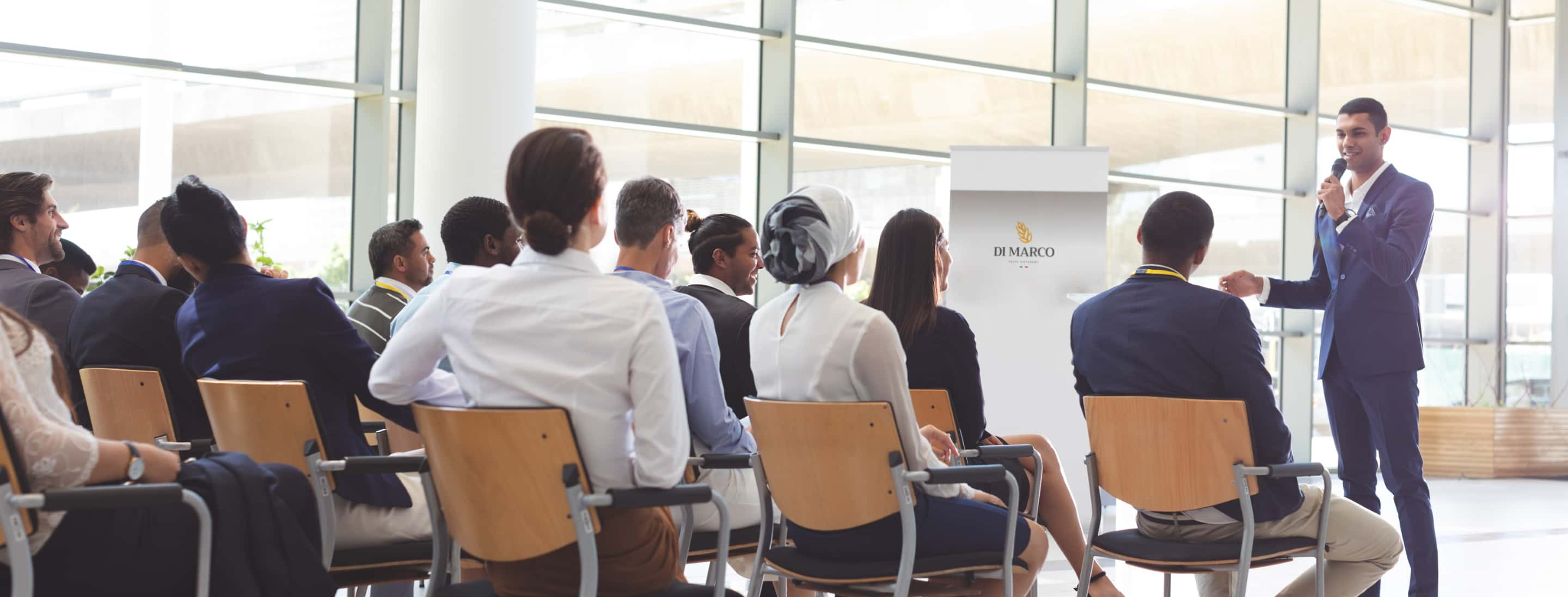 Photo of a classroom with a speaker speaking into a microphone and an audience seated to listen.