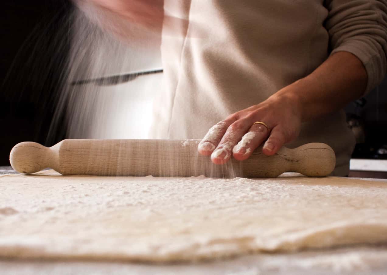 Dettaglio della mani di un cuoco mentre stende un impasto con il mattarello e cosparge della farina